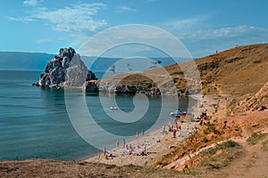 Cape Shamanka rock in clear blue Lake Baikal among grassy steppess, sand coast against the background of mountains