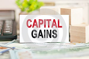 CAPITAL GAINS text on white paper, on the background of bills and wooden blocks, business concept