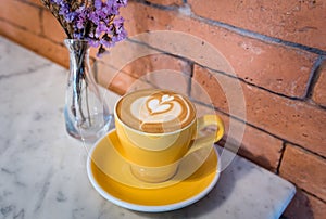 Cappuccino coffee of a heart shaped in a yellow cup near purple