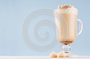Cappuccino coffee with whipped cream, cocoa powder and biscuits in transparent glass with handle on pastel blue wall.
