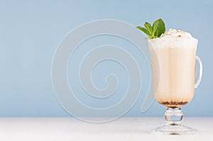 Cappuccino coffee with whipped cream, green mint, cocoa powder in  transparent glass with handles on pastel blue wall.