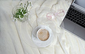 Cappuccino coffee in white cup and saucer, beautiful soft beige plaid, spring flowers, laptop, concept of holiday, mother`s day,