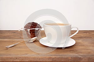 Cappuccino coffee in a white cup and saucer and chocolate muffin in paper on an old wooden table, concept of sweet life