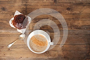 Cappuccino coffee in a white cup and saucer and chocolate muffin in paper on an old wooden table, concept of sweet life, top view