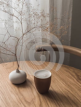 Cappuccino cup, vase with dried flowers on a wooden round table