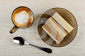 Cappuccino in cup, wafers in brown saucer, spoon on wooden table. Top view