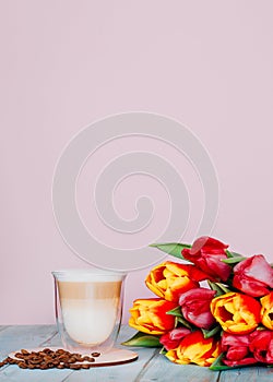 Cappuccino in a glass with double walls and roasted coffee beans with a bouquet of tulips. Feminine pink background with