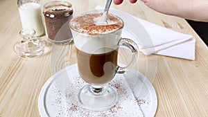 Cappuccino, latte with froth milk in cup. Hand stirs spoon of coffee in tall glass, scoops up whipped cream, sprinkled with ground
