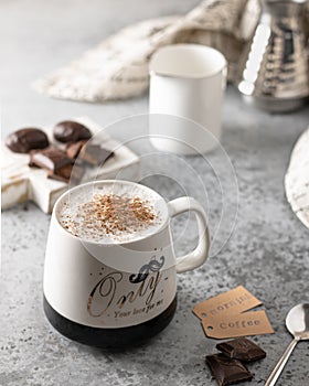 Cappuccino in a white porcelain mug with milk foam, decorated with ground cinnamon and sugar on a gray textured table.