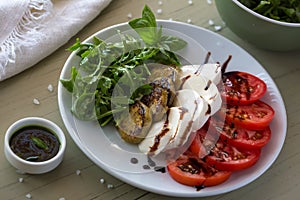 Caprese salad with mozzarella, tomato, basil and grilled eggplant on white plate. Top view on background