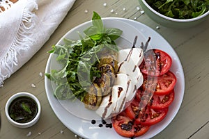 Caprese salad with mozzarella, tomato, basil and grilled eggplant on white plate. Top view on background