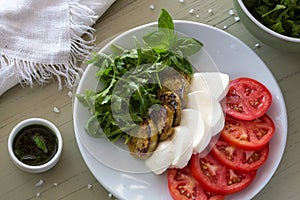 Caprese salad with mozzarella, tomato, basil and grilled eggplant on white plate. Top view on background