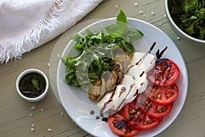 Caprese salad with mozzarella, tomato, basil and grilled eggplant on white plate. Top view on background