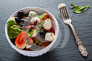 Caprese salad, small mozzarella cheese, fresh green leaves, black olives and cherry tomatoes in white vintage bowl on stone