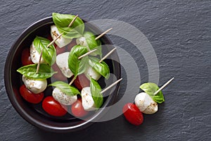 Caprese sticks with mozzarella, tomatoes and basil leaves in bowl on black background