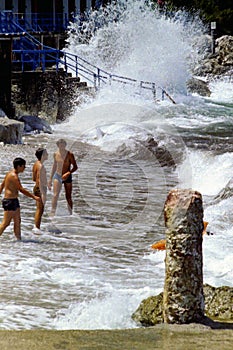 CAPRI, ITALY, JULY 1969 - Three boys defy the waves during a swell between the pebbles, the rocks and a Roman column on the beach.