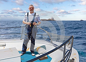 The captain of a ship stands at the bow with binoculars and looks into the camera