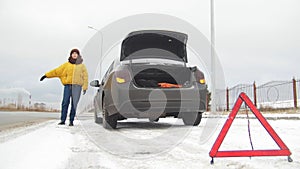 A car accident. A young woman standing by a broken car. Catching a car for help. An emergency sign