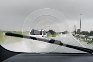 A car appearing clearly as a windscreen is wiped of rain by a wiper. Spray from cars can be seen on the wet road.