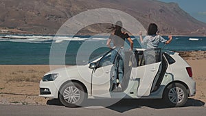 Car on beautiful sandy shore. Two young attractive girls running towards the car in slow motion. Girls then admiring