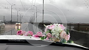 A car with a bouquet on the hood passes a bridge in a rainy city. wipers clean the windshield. the road and rain are