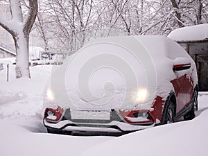 A car with burning headlights is buried under the snow after a snowstorm in a residential area