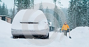 Car covered with snow as a huge snowdrift on the countryside forest home driveway. Smiling man with a shovel going to remove snow