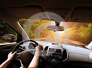 Car dashboard with driver`s hands on the steering wheel and rear view mirrors on a road in motion with trees against sky with