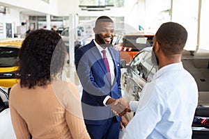 Car Dealership Concept. Salesperson Handshaking With Young Black Customers Couple In Showroom
