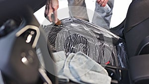 Car detailing concept, young man cleaning vehicle seats with a rag and a brush in car studio, car care
