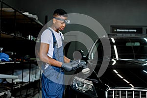 Car detailing and polishing concept. Professional black male car service worker in uniform, holding in hands orbital