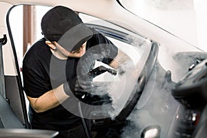 Car detailing, steam cleaning concept. Handsome man in black t-shirt and cap, worker of car wash center, cleaning car