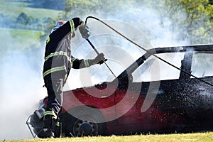 Car fire. A firefighter extinguishes a car fire with foam. Red car on fire, white smoke and firefighter. Accident in the meadow