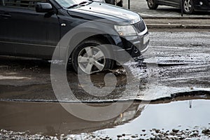 The car goes without reducing speed on a bad road with asphalt in pits and potholes, hitting the wheel in a puddle, spraying melt
