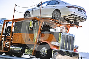 Car hauler big rig orange semi truck transporting cars on the modular hydraulic semi trailer moving on the highway road