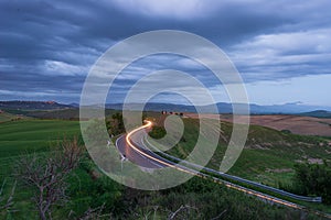 Car light trail on winding road in the unique hill landscape of Tuscany, Italy. Dramatic sunset sky at twilight, illuminated