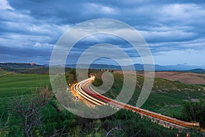 Car light trail on winding road in the unique hill landscape of Tuscany, Italy. Dramatic sunset sky at twilight, illuminated