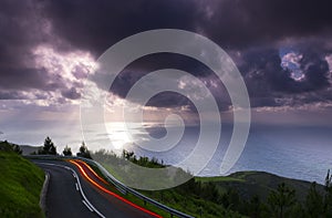 Car lights on the mountain road with clouds and sun over the sea at sunset, mount Jaizkibel