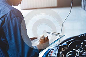 Car mechanic holding clipboard and checking to maintenance vehicle by customer claim order in auto repair shop garage. Engine