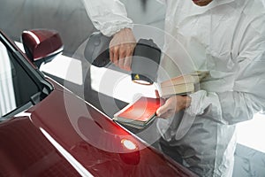 Car painter man determines the correct tone of a vehicle body colour using a special colorist`s lamp.