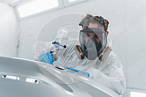 Car painter in protective clothes and mask painting and varnish automobile bumper in chamber.