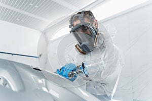 Car painter in protective clothes and mask painting and varnish automobile element in chamber.