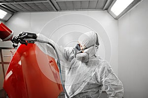 Car painter spraying red paint with spray gun on vehicle element in paint chamber.