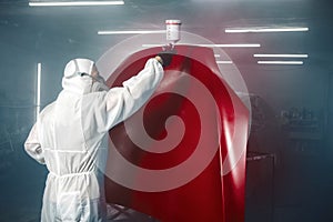 Car painter spraying red paint with spray gun on vehicle element in paint chamber.