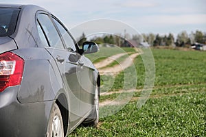 The car is parked in the field. The car is driving along the rural road to the house. The car is gray in the meadow in front of