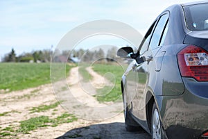The car is parked in the field. The car is driving along the rural road to the house. The car is gray in the meadow in front of