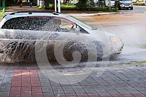 The car passes a pedestrian crossing, flooded with water after rain. Cloud splashing water. Sunny day. Reflection in a