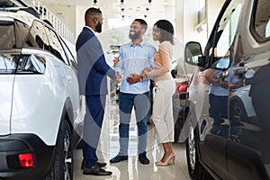 Car Purchase. Happy Customers Couple Handshaking With Manager In Auto Salon