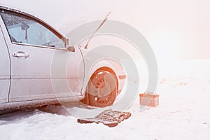 A car with a raised hood on a background of a snowy field, tools and a discharged car battery near the car, the concept of a car