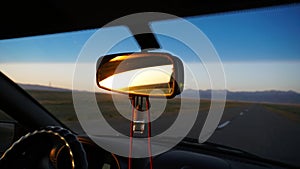 Car rear view mirror. Displays orange dawn. Visible road, hills and steppe.